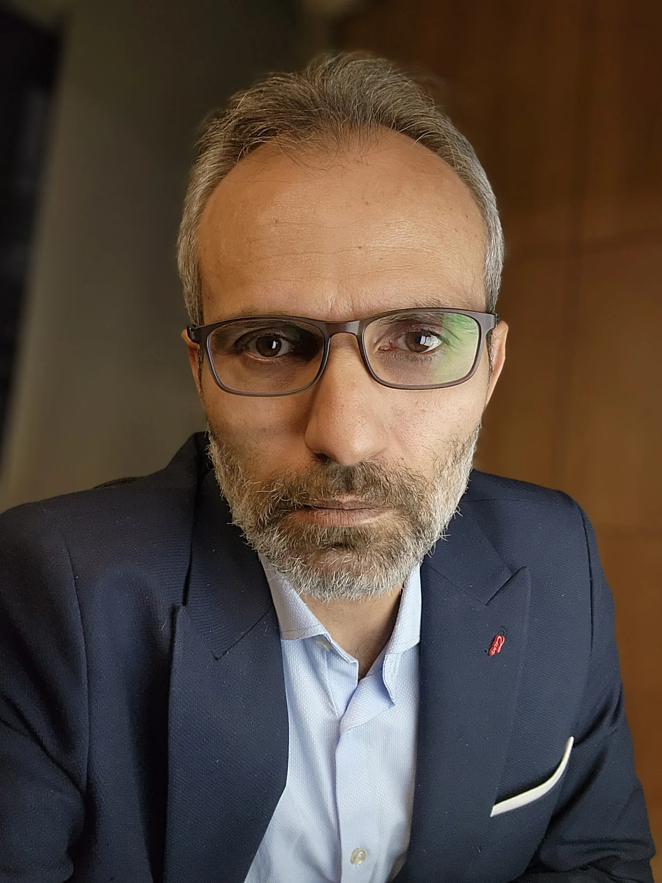 Galip Arduç, koyu lacivert ceket ve gözlükle sıcak tonlu bir iç mekânda kameraya bakıyor.