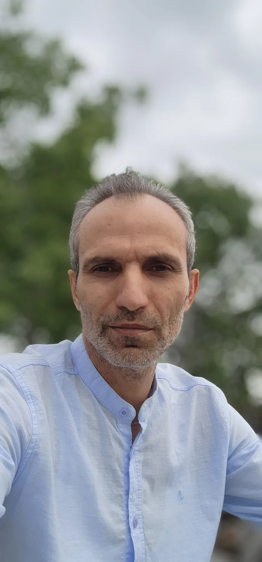Galip Arduç'un açık mavi hâkim yaka gömleğiyle, yeşil ağaçların önünde, doğal ışıkta ve sakin bir ifadeyle çekilmiş açık hava portresi.