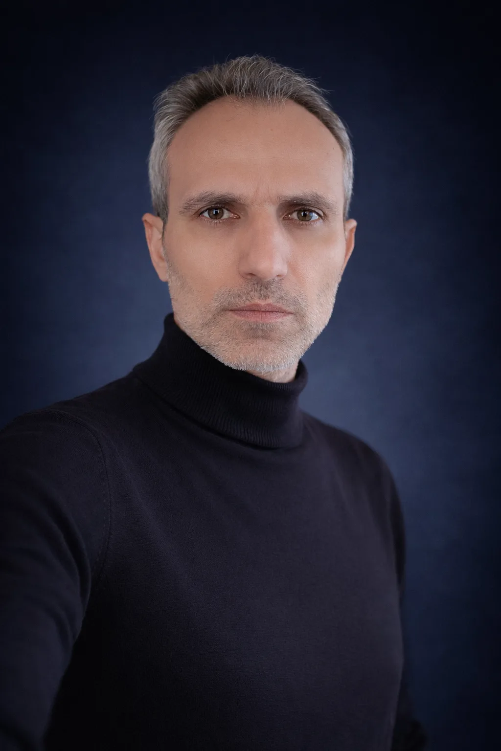 Galip Arduç’un koyu tonlu stüdyo atmosferinde siyah kazakla çekilmiş ciddi ve modern portresi.