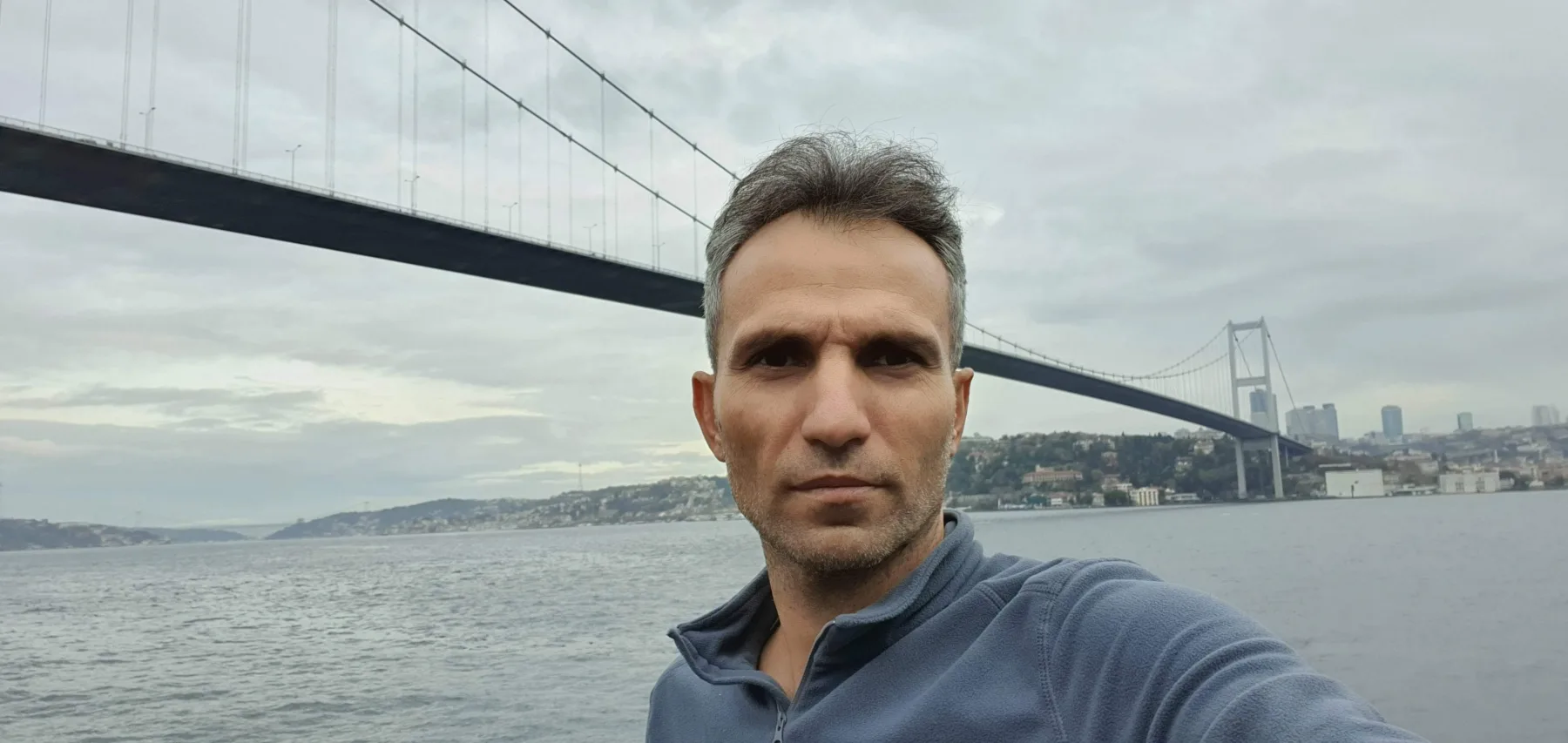 Galip Arduç’un İstanbul Boğazı ve köprü manzarası önünde, sakin ve kararlı duruşuyla çekilmiş açık hava portresi.