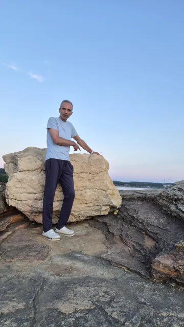 Galip Arduç'un Kefken Pembe Kayalıklar'da açık mavi tişörtüyle büyük kayanın yanında ayakta durduğu, doğal manzara ve açık gökyüzü eşliğinde çekilmiş fotoğrafı.