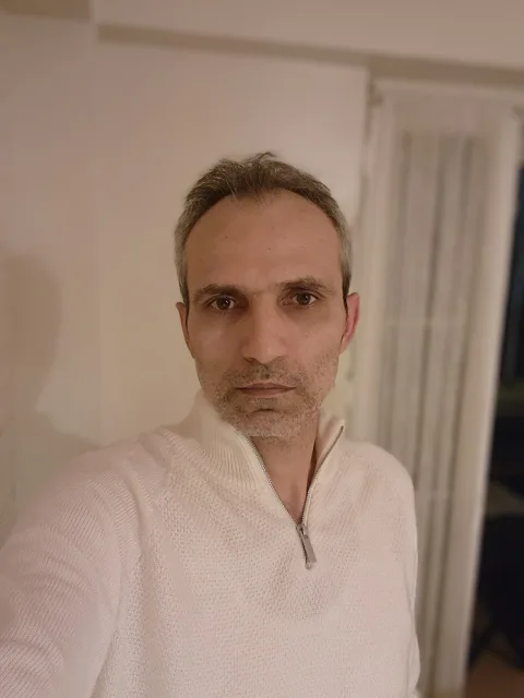 Galip Arduç, açık renk fermuarlı triko ile iç mekânda kameraya doğrudan bakıyor.