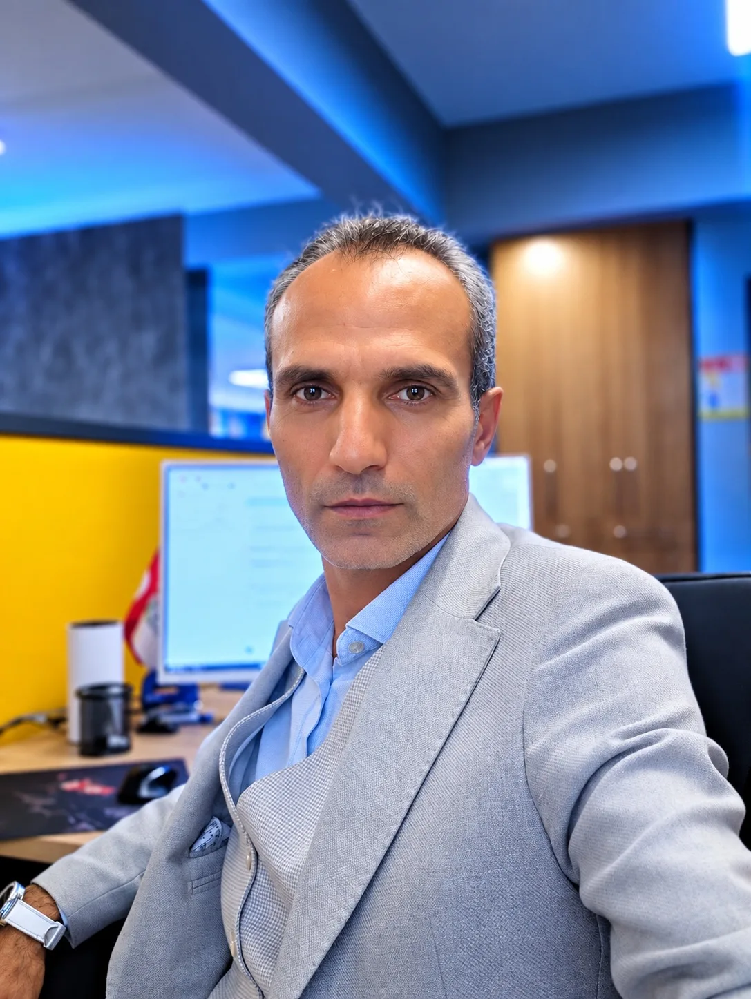 Galip Arduç, sarı bölmeli ve mavi ışıklarla aydınlatılmış bir ofiste açık gri takım elbiseyle kameraya bakıyor.