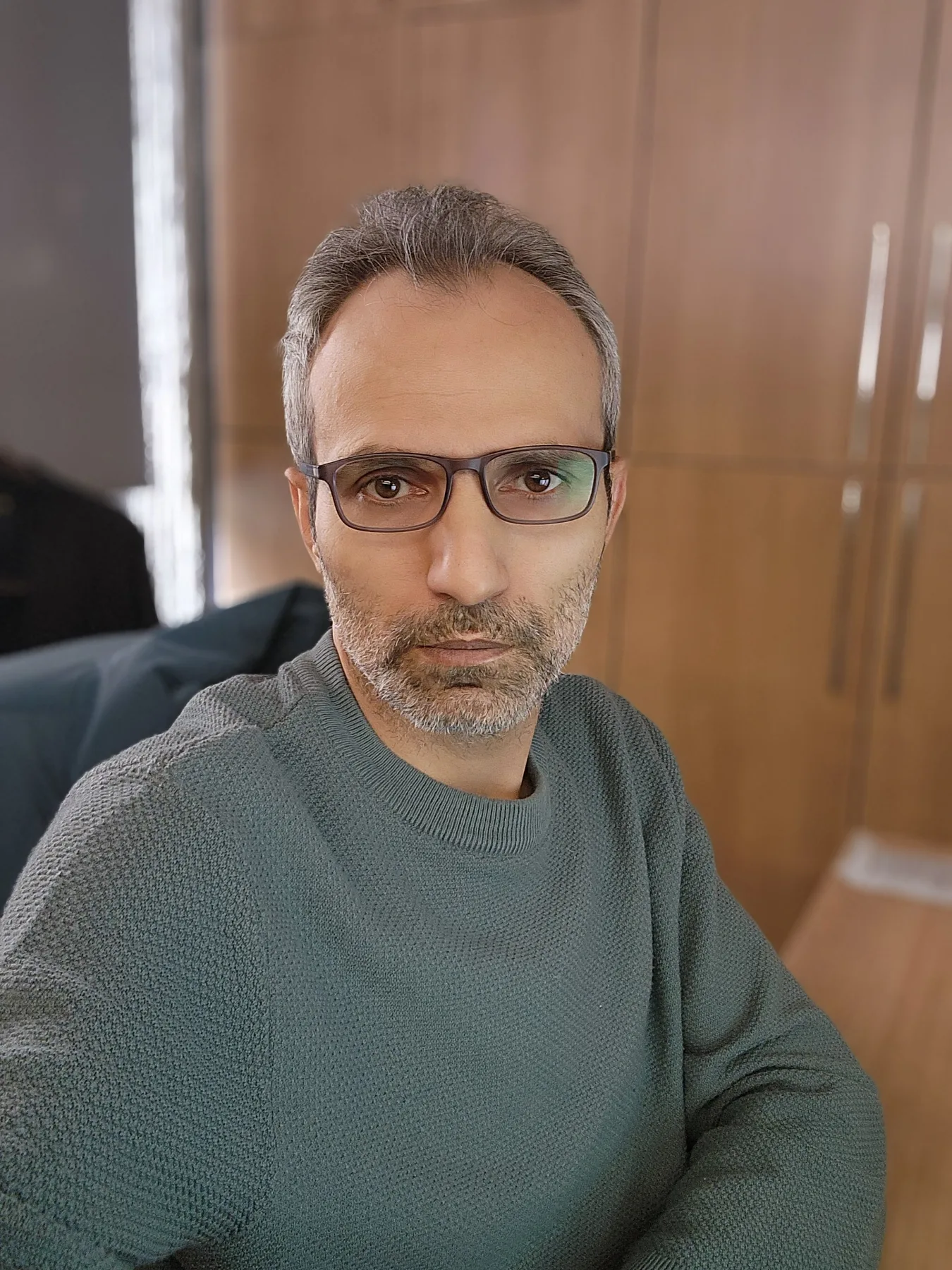 Galip Arduç, ahşap dolapların önünde gri kazak ve gözlükle otururken kameraya bakıyor.