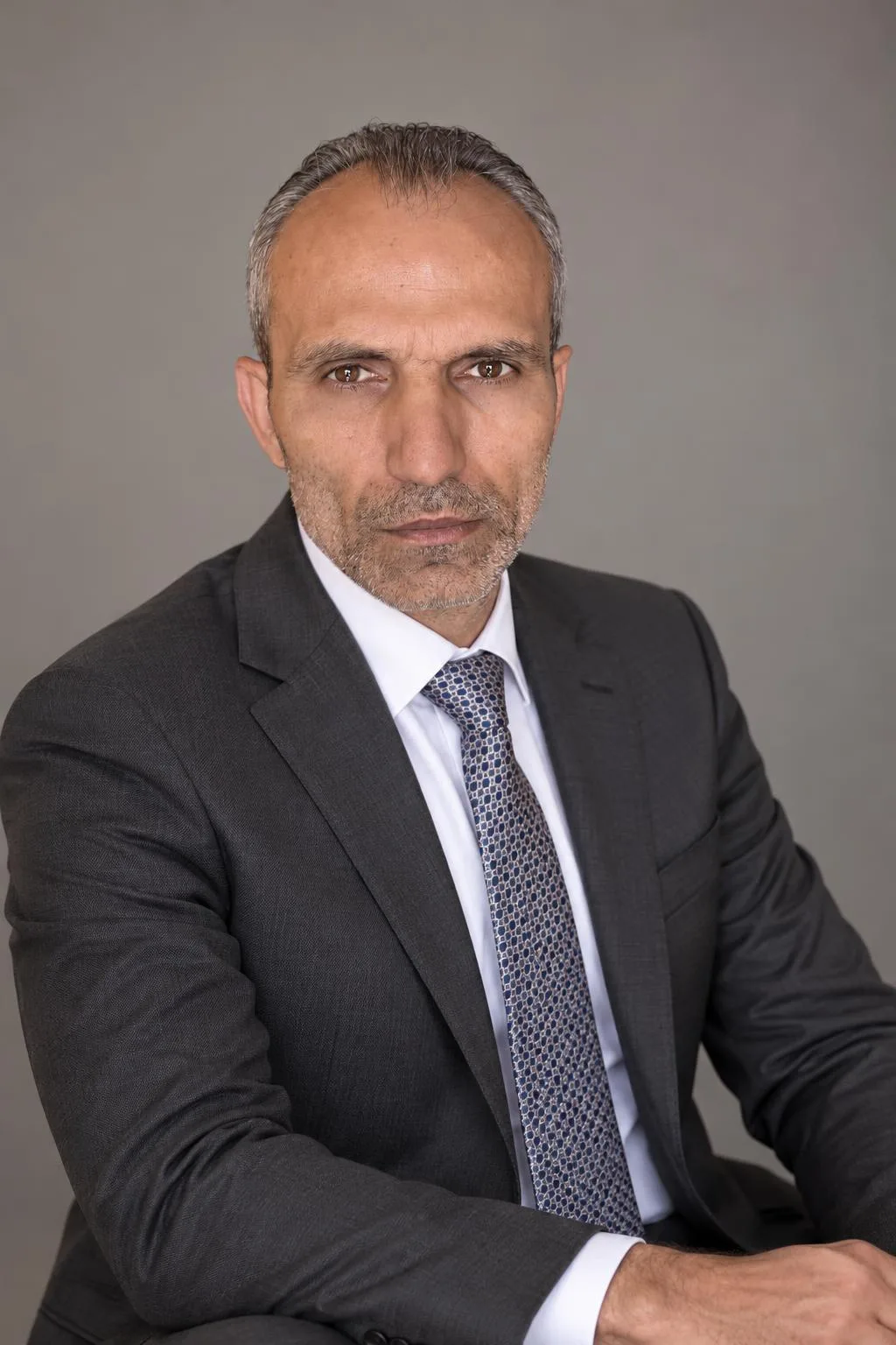Galip Arduç, gri fon önünde koyu renk takım elbise ve desenli kravatla otururken kameraya bakıyor.