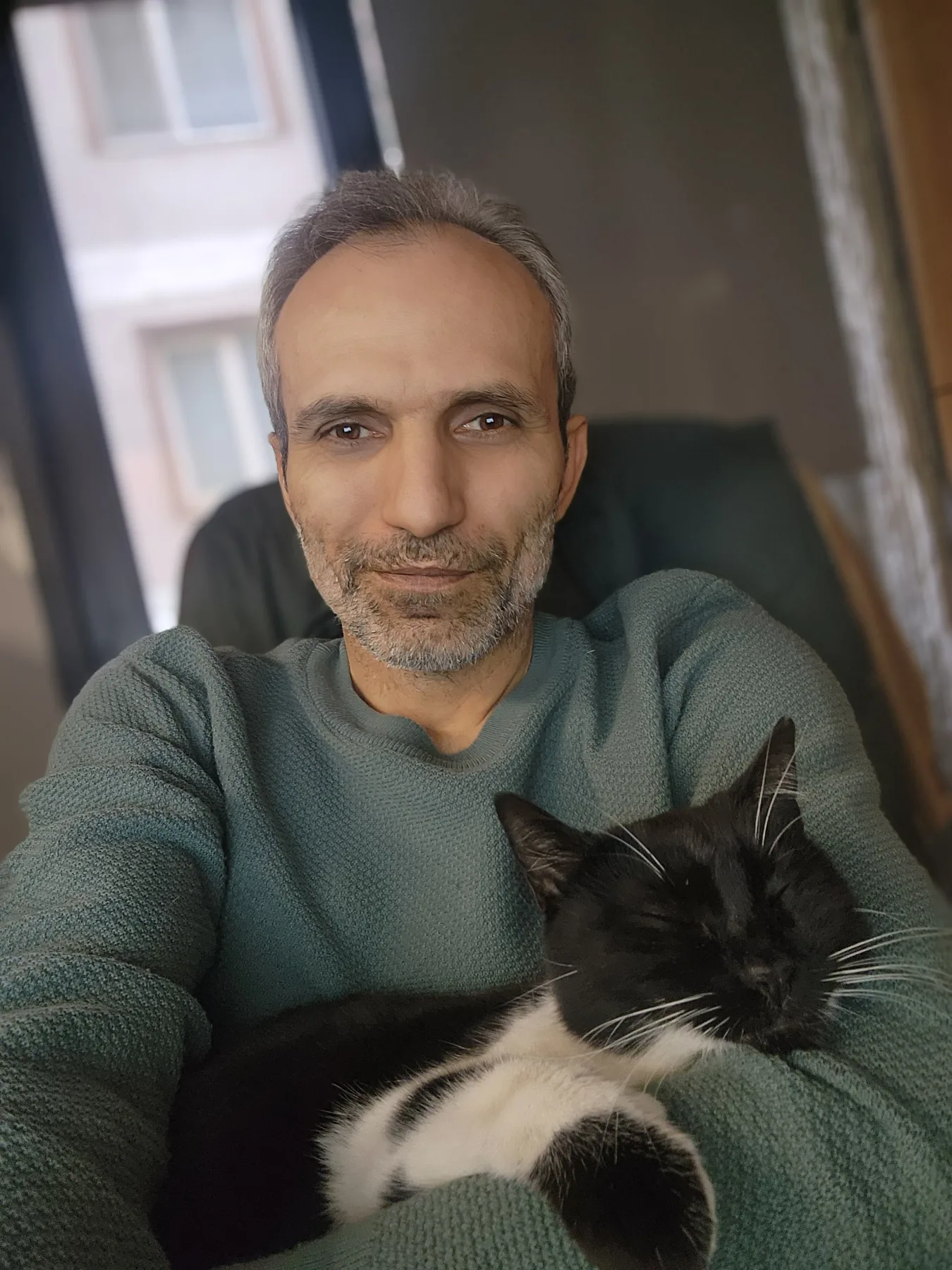 Galip Arduç’un fotoğrafında, yeşil kazaklı bir kişi kucağında siyah beyaz bir kediyle oturuyor.