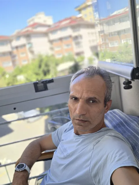 Galip Arduç, açık renk tişörtüyle camlı balkonda otururken kameraya bakıyor.
