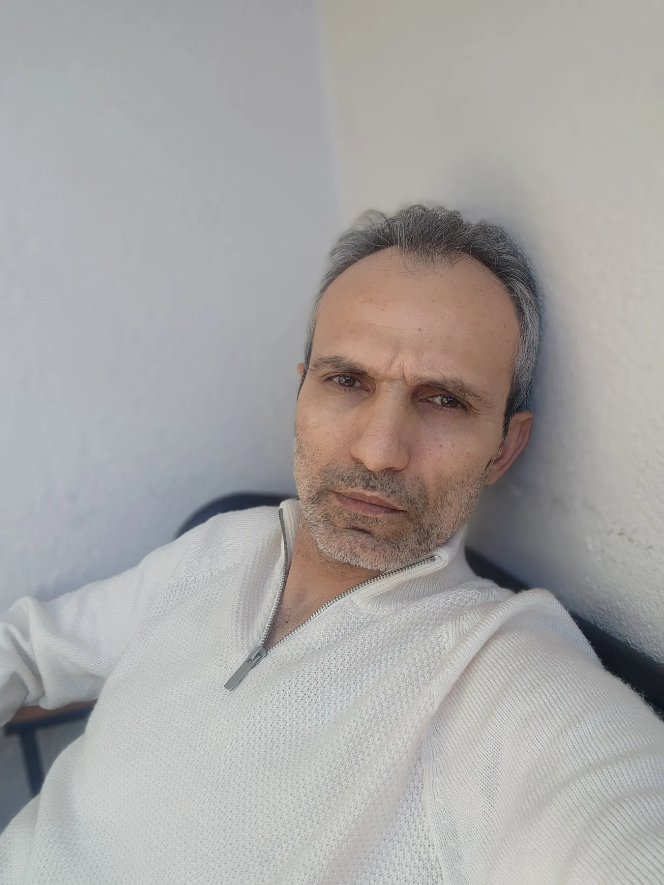 Galip Arduç, açık renk fermuarlı kazağıyla açık tonlu bir duvarın yanında otururken kameraya bakıyor.