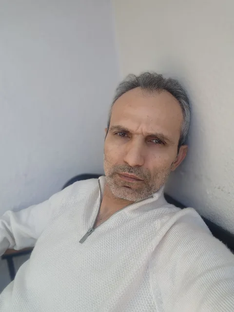 Galip Arduç, açık renk fermuarlı kazağıyla açık tonlu bir duvarın yanında otururken kameraya bakıyor.