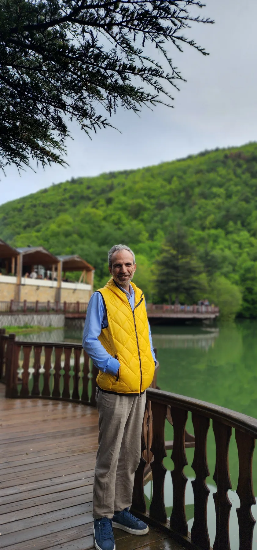 Galip Arduç, yeşil yamaçlar ve sakin su manzarası önünde ahşap bir terasta duruyor.
