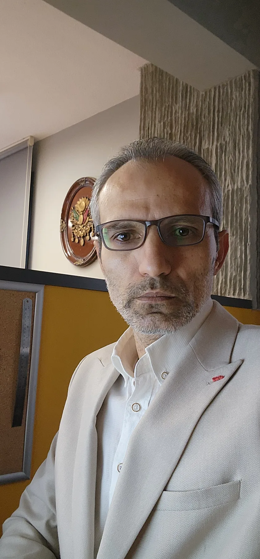 Galip Arduç'un krem rengi ceketi, beyaz gömleği ve siyah çerçeveli gözlüğüyle iç mekânda çekilmiş, ciddi ve kendinden emin duruşunu yansıtan profesyonel portresi.